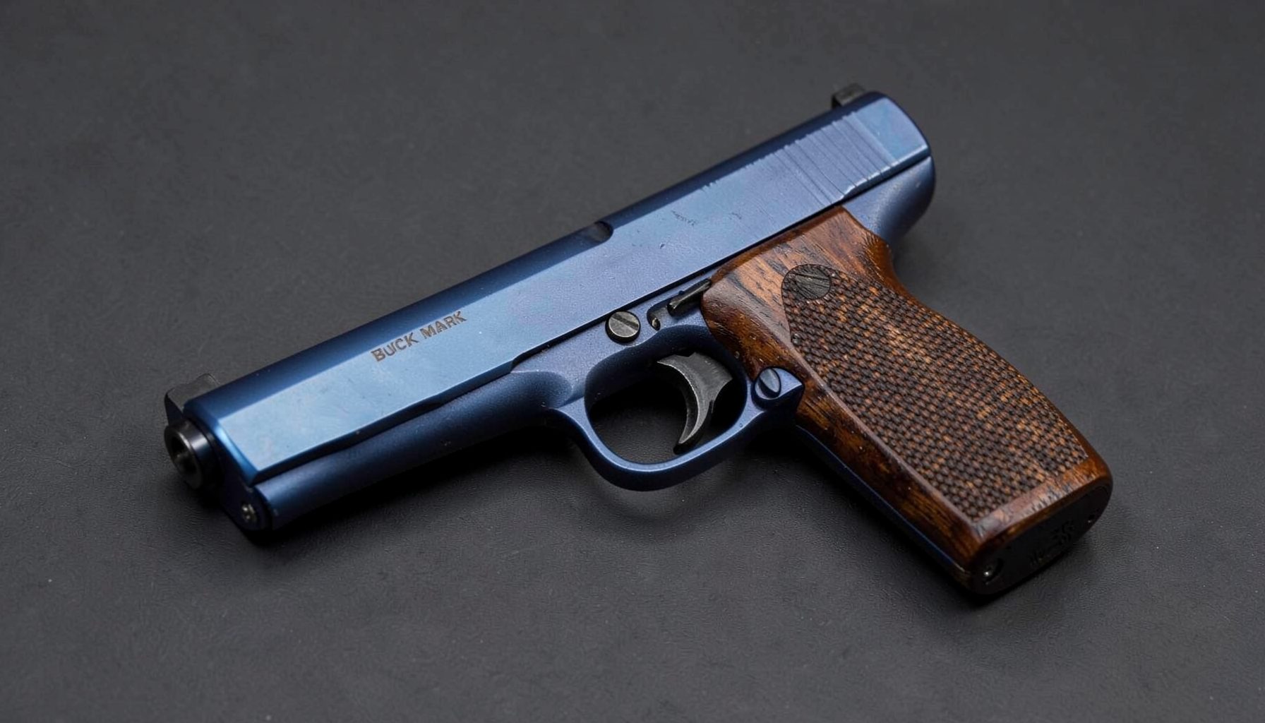 A Browning Buck Mark pistol with a blued steel slide and checkered wood grip panels, resting on a dark matte surface under soft studio lighting.