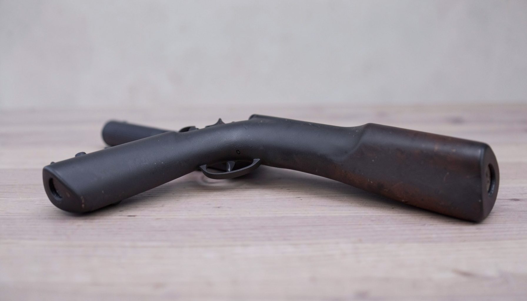 A muted charcoal-grey and soft purple still-life arrangement of a rifle stock and shotgun barrel resting on a plain wooden surface, calm and understated in mood